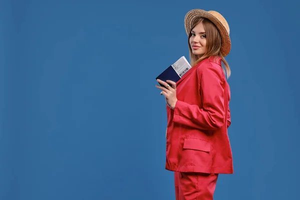 Blonde woman in straw hat, white blouse and red pantsuit. She smiling, holding passport and ticket while posing sideways on blue background. Close-up — 스톡 사진
