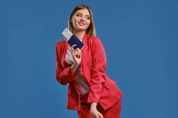 Блондинка в белой блузке и красном брючном костюме. Она улыбается, держа в руках паспорт и билет, позируя на фоне синей студии. Крупный план — стоковое фото