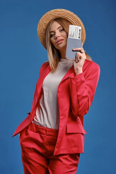 Blonde lady in straw hat, white blouse and red pantsuit. She is showing passport and ticket while posing on blue studio background. Close-up — 스톡 사진