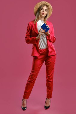 Blonde lady in straw hat, white blouse, red pantsuit and high black heels. She holding passport and ticket, posing on pink background. Full length