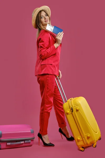 Attractive blonde girl in straw hat, red pantsuit, high black heels. She smiling, holding yellow suitcase, passport and ticket, pink background — Stock Fotó