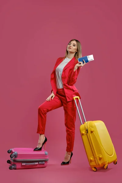 Blonde woman in red pantsuit, white blouse, black heels. She put her leg on suitcase, yellow one nearby, holding passport and ticket, pink background — Stockfoto