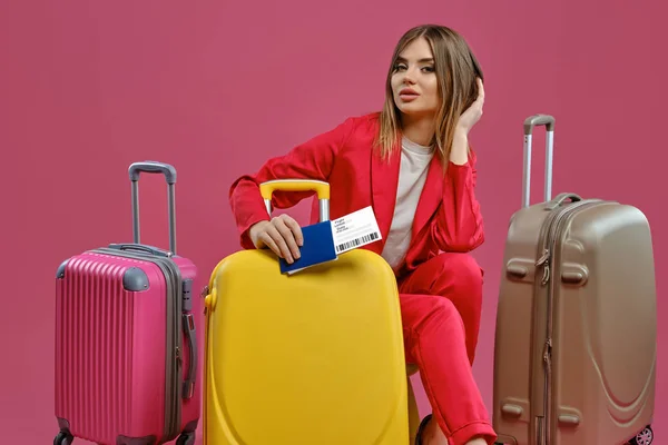 Блондинка в красном брючном костюме, белая блузка. Она трогает волосы, сидит среди разноцветных чемоданов, держит паспорт и билет, розовый фон — стоковое фото