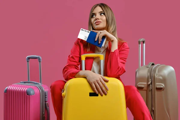 Blonde girl in red pantsuit, white blouse. She looking thoughtful, sitting among colorful suitcases, holding passport and ticket, pink background — Stockfoto