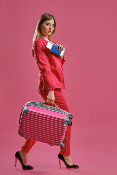 Blonde girl in red pantsuit, high black heels. She is carrying suitcase, holding passport and ticket, pink background. Travelling concept — Stok fotoğraf