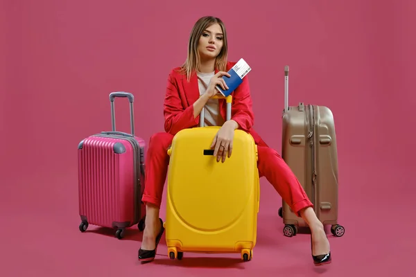 Senhora loira de fato vermelho, blusa branca, salto alto e preto. Ela sentada entre três malas coloridas, segurando passaporte e bilhete, fundo rosa — Fotografia de Stock