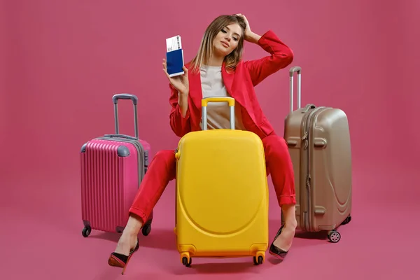 Блондинка в червоному костюмі, біла блузка, чорні підбори. Вона посміхається, сидить серед різнокольорових валіз, тримає паспорт і квиток, рожевий фон — стокове фото