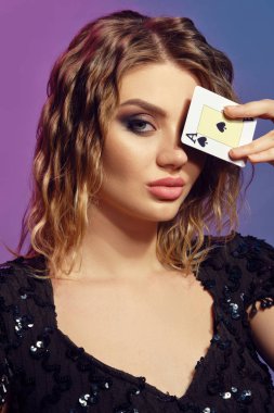 Blonde lady in black sequin dress has covered her eye with ace of spades, posing on colorful studio background. Gambling, poker, casino. Close-up.