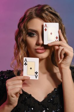 Brown-haired woman in black shiny dress showing two playing cards, posing on colorful background. Gambling entertainment, poker, casino. Close-up.
