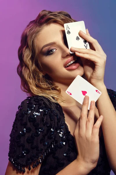 Donna dai capelli castani in abito nero lucido sorride, mostrando due carte da gioco, in posa su sfondo colorato. Gioco d'azzardo, poker, casinò. Primo piano . — Foto Stock