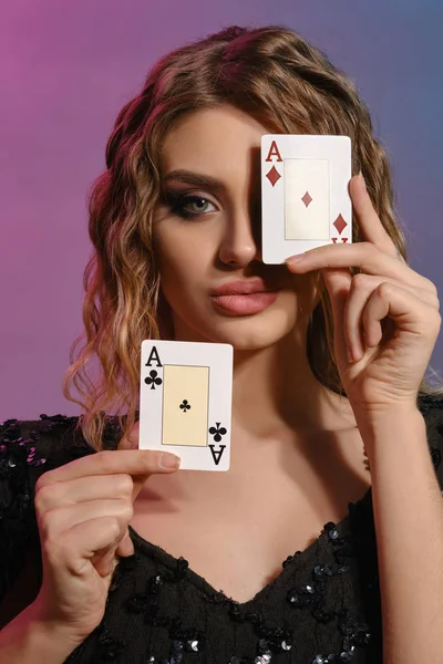 Mujer de cabello castaño en vestido negro brillante que muestra dos cartas de juego, posando sobre un fondo colorido. Entretenimiento, póquer, casino. Primer plano . — Foto de Stock