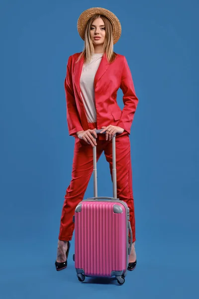 Blonde lady in straw hat, white blouse, red pantsuit, high black heels. She is leaning on a handle of pink suitcase while posing on blue background — Stock fotografie