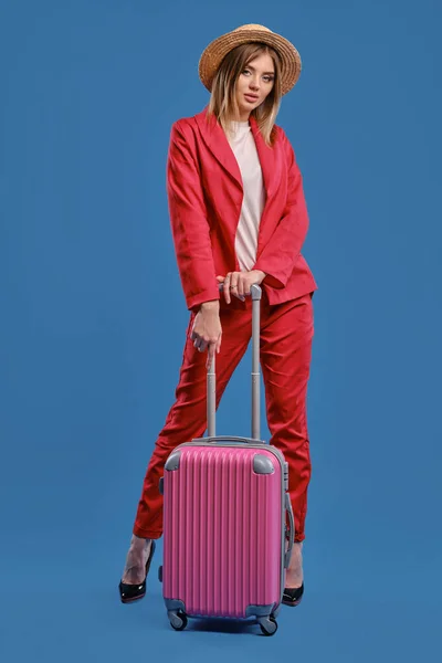 Blonde model in straw hat, white blouse, red pantsuit, high black heels. She is leaning on a handle of pink suitcase while posing on blue background — Stock fotografie