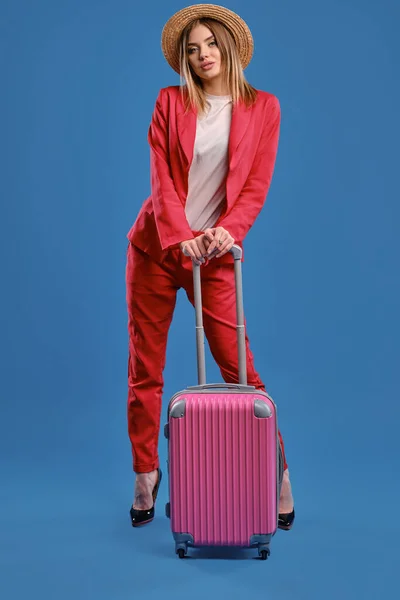 Blonde woman in straw hat, white blouse, red pantsuit, high black heels. She is leaning on a handle of pink suitcase while posing on blue background — Stok fotoğraf
