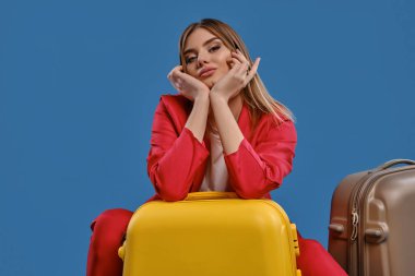 Blonde lady in white blouse and red pantsuit. She leaning on yellow suitcase, gray one nearby, posing on blue studio background. Close-up