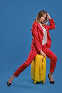 Blonde girl in white blouse, red pantsuit, high black heels. She touching hair, sitting on yellow suitcase, posing on blue background. Full length
