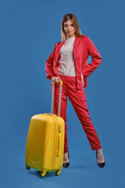 Blonde lady in red pantsuit, white blouse and high black heels. She is holding yellow suitcase, posing against blue studio background. Full length