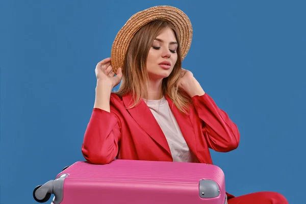 Blonde model in straw hat, white blouse, red pantsuit. Sitting leaning on pink suitcase, posing on blue background. Travelling concept. Close-up — Stock fotografie