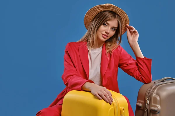 Blonde model in straw hat, white blouse, red pantsuit. She sitting leaning on yellow suitcase, posing on blue background. Close-up, copy space — Stock fotografie