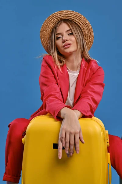 Blonde female in straw hat, white blouse, red pantsuit. She sitting leaning on yellow suitcase, posing on blue background. Close-up, copy space — Zdjęcie stockowe