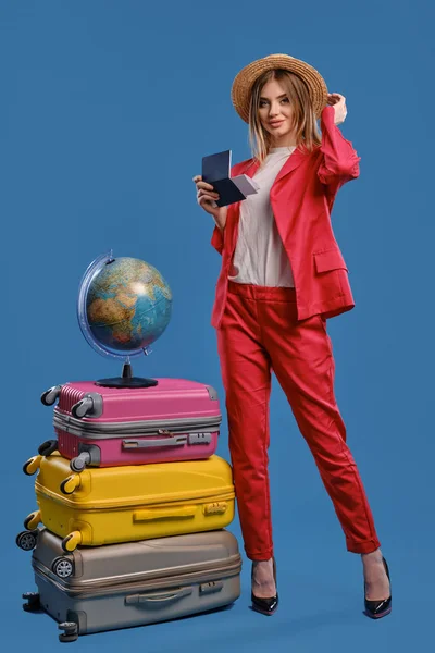 Модель в шляпе, белая блузка, красный брючный костюм, черные каблуки. Улыбается, держит паспорт и билет, позирует на синем фоне. Глобус на чемоданах — стоковое фото