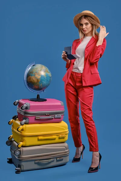 Female in straw hat, white blouse, red pantsuit, black heels. Holding passport and ticket, posing on blue background. Globe is on top of suitcases — Stock fotografie