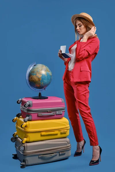 Girl in straw hat, white blouse, red pantsuit, black heels. Holding passport and ticket, posing on blue background. Globe is on top of suitcases — Stock fotografie