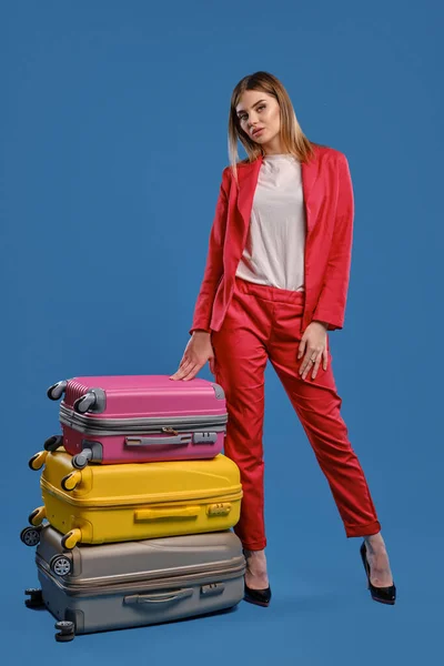 Woman in white blouse, red pantsuit, high black heels. She is touching stacked colorful suitcases, posing on blue studio background. Full length — Stock fotografie