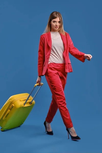 Blonde female in white blouse, red pantsuit and high black heels. She carrying yellow suitcase by handle while walking on blue background. Full length — Stock fotografie