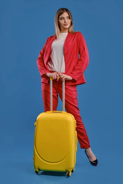 Blonde female in red pantsuit, white blouse and high black heels. She is holding yellow suitcase, posing against blue studio background. Full length — Stock fotografie