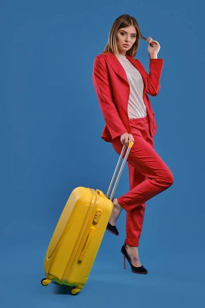 Blonde woman in red pantsuit, white blouse and high black heels. She is holding yellow suitcase, posing against blue studio background. Full length — Stok fotoğraf