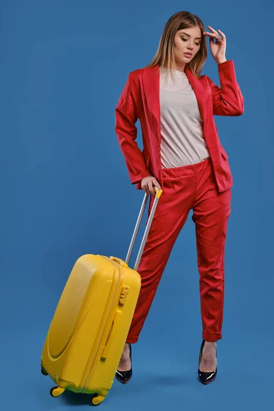 Blonde model in red pantsuit, white blouse and high black heels. She is holding yellow suitcase, posing against blue studio background. Full length — Stock fotografie