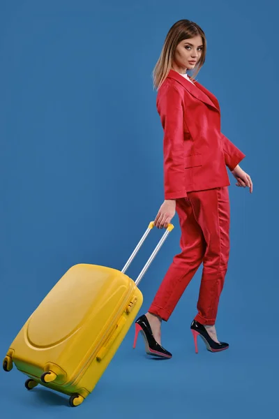 Chica rubia en traje pantalón rojo, tacones negros altos. Ella está posando de nuevo a la cámara con la maleta amarilla sobre fondo azul del estudio. Longitud completa — Foto de Stock
