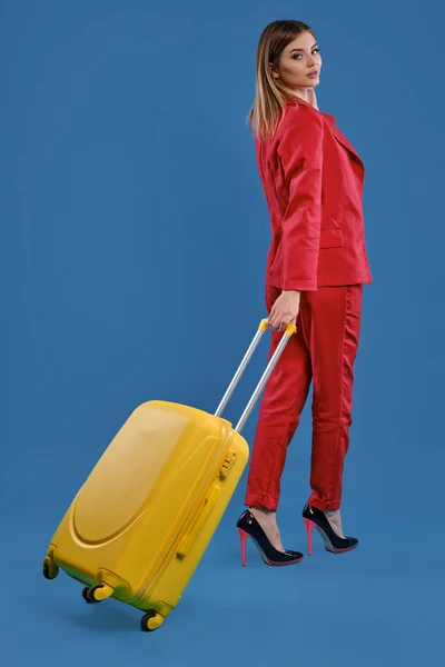 Blonde female in red pantsuit, high black heels. She is posing back to the camera with yellow suitcase on blue studio background. Full length — Stok fotoğraf