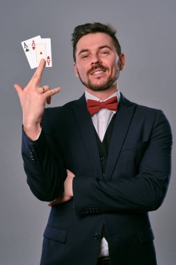 Man in black classic suit and red bow-tie showing two playing cards while posing against gray studio background. Gambling, poker, casino. Close-up.