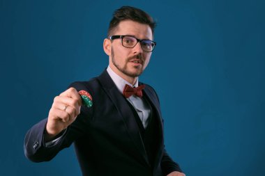 Man in black classic suit, red bow-tie, glases is showing two colored chips, posing on blue studio background. Gambling, poker, casino. Close-up.