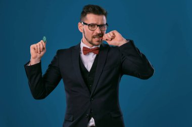 Man in black classic suit, red bow-tie, glases is showing two colored chips, posing on blue studio background. Gambling, poker, casino. Close-up.