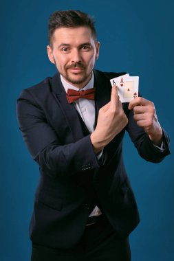Man in black classic suit and red bow-tie showing two playing cards while posing against blue studio background. Gambling, poker, casino. Close-up.