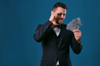 Man in black classic suit and red bow-tie showing one chip and some dollar bills, posing on blue background. Gambling, poker, casino. Close-up.