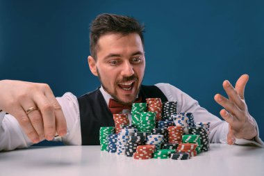 Man in black vest and shirt sitting at white table with stacks of chips on it, posing on blue studio background. Gambling, poker, casino. Close-up.
