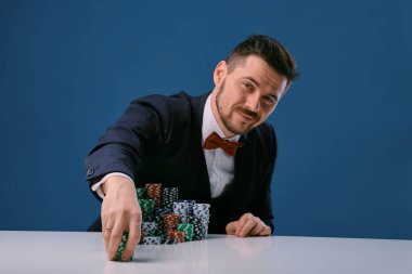 Man in black suit is sitting at white table with colored stacks of chips on it, posing on blue studio background. Gambling, poker, casino. Close-up.