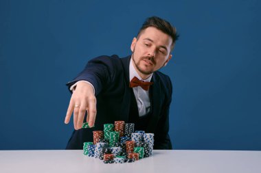 Man in black suit is sitting at white table with colored stacks of chips on it, posing on blue studio background. Gambling, poker, casino. Close-up.
