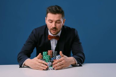 Man in black suit is sitting at white table with colored stacks of chips on it, posing on blue studio background. Gambling, poker, casino. Close-up.