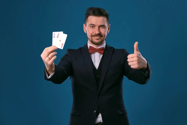 Man in black classic suit and red bow-tie showing two playing cards while posing against blue studio background. Gambling, poker, casino. Close-up. — Stockfoto