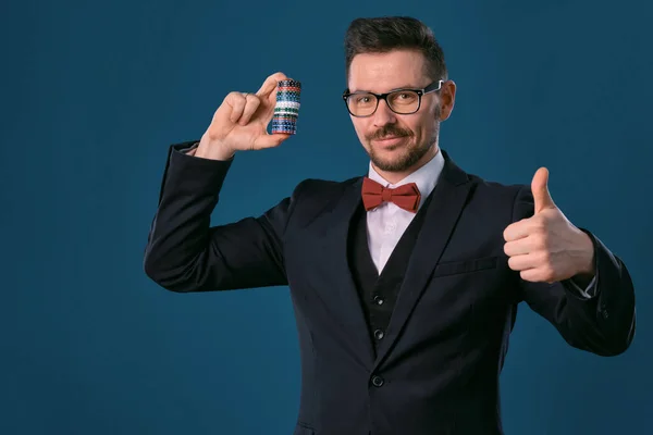 Man in black .ic suit, red bow-tie, glases is showing some colored chips, posing on blue studio background. Gambling, poker, casino. Close-up. — Stock Photo, Image