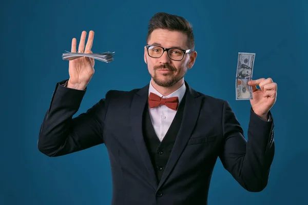 Man in black classic suit, red bow-tie, glases is holding some dollar bills, posing on gray studio background. Gambling, poker, casino. Close-up. — ストック写真