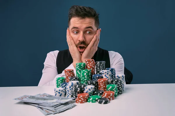 Hombre en chaleco negro y camisa sentado en la mesa blanca con montones de fichas y dinero en efectivo en ella, posando sobre fondo azul. Apuestas, póquer, casino. Primer plano . —  Fotos de Stock