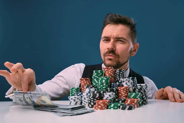 Man in black vest and shirt sitting at white table with stacks of chips and cash on it, posing on blue background. Gambling, poker, casino. Close-up. — Stockfoto