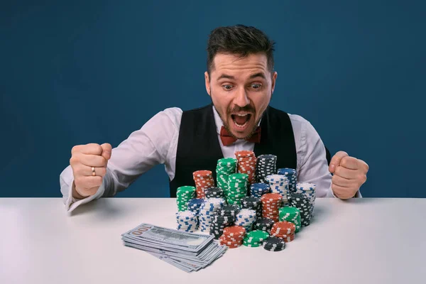 Man in black vest and shirt sitting at white table with stacks of chips and cash on it, posing on blue background. Gambling, poker, casino. Close-up. — Stockfoto