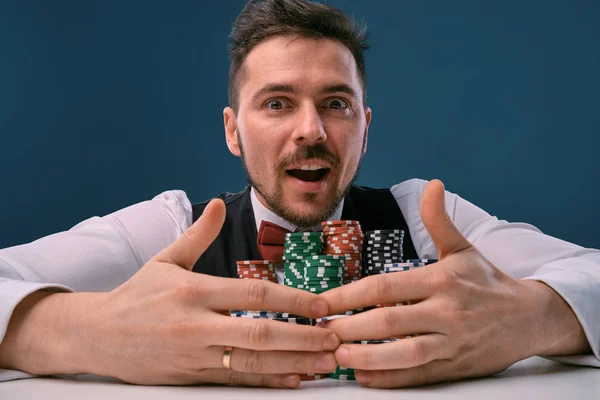 Чоловік в чорному жилеті і сорочці, сидячи за білим столом зі стеками чіпсів на ньому, позує на синьому студійному фоні. Азартні ігри, покер, казино. Крупним планом . — стокове фото